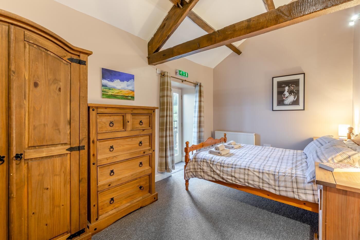Een slaapkamer met een houten dressoir, een bed met geruit beddengoed, een ingelijst schilderij en een raam met gordijnen.