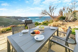 An outdoor dining area with a view of the ocean, a grill, and a table set with plates and food. - 2 Bed in Sennen (oc-t30543) (Sennen)