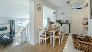 A modern kitchen with white appliances, a dining area with a wooden table and white chairs, and a living area with a grey sofa and a TV.