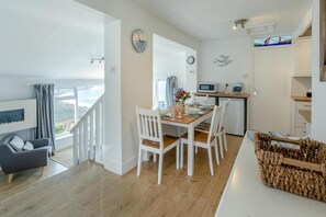 A modern kitchen with white appliances, a dining area with a wooden table and white chairs, and a living area with a grey sofa and a TV. - 2 Bed in Sennen (oc-t30543) (Sennen)