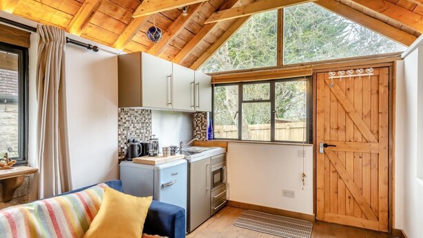 A compact kitchen with a small refrigerator, a sink, and a window. There's a wooden door with heart-shaped decorations and a window with a view of trees. - 1 Bed in Swanage (oc-l30493) (Harman's Cross)