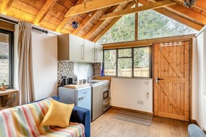Una cocina compacta con un refrigerador pequeño, un lavamanos y una ventana. Hay una puerta de madera con detalles de corazones y una ventana con vista a árboles.