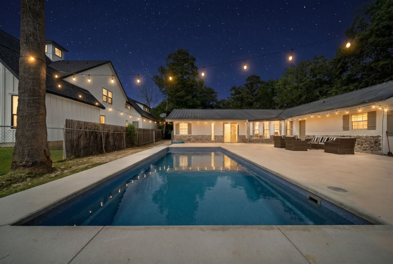 A swimming pool with a night sky view, surrounded by houses and string lights.