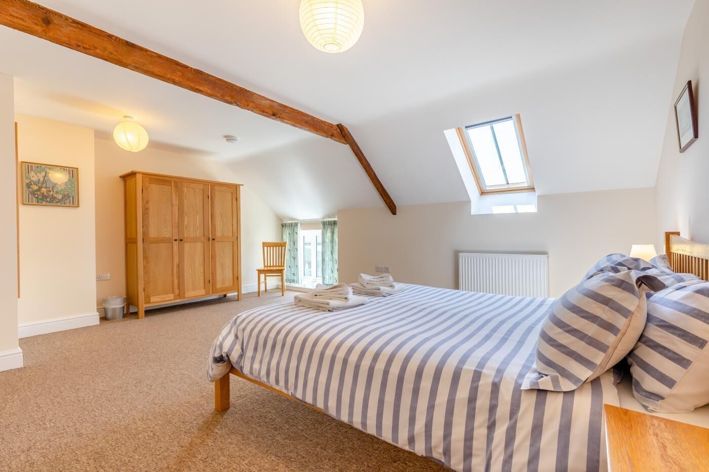 Ein Schlafzimmer mit schrägverdeckter Decke, einem Bett mit gestreifter Bettwäsche, einem Holzschrank, einem Stuhl und einem Fenster, das natürliches Licht hereinlässt.