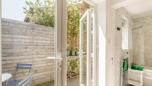 A room with a wooden fence, a glass door leading to a garden, a small table with chairs, and a bathroom with a tiled floor and a green towel.