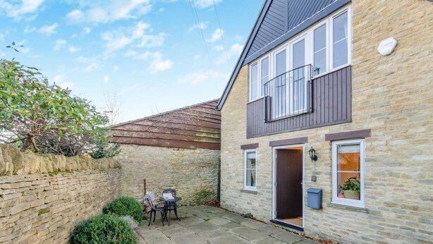 Une maison en pierre avec un balcon, une porte et un petit espace extérieur aménagé pour s’asseoir.
