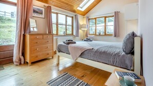 Quarto com teto de madeira, cama com cobertor cinzento, penteadeira e janela com cortinas.
