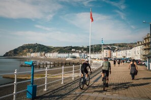 A coastal promenade with cyclists and pedestrians, a beach, and historic buildings in the background.