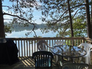 A wooden deck with a glass table and chairs overlooking a calm lake, surrounded by trees.