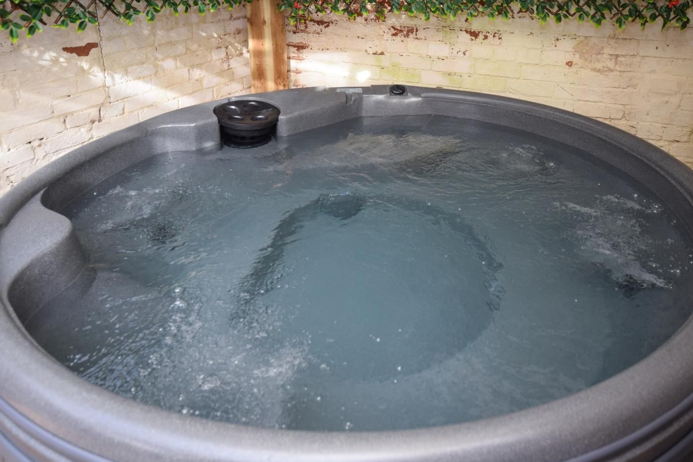 A hot tub with a black jet nozzle, surrounded by a brick wall and greenery.