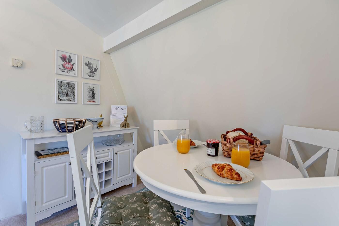 Un coin repas avec une table ronde, des chaises, un panier de pain, une assiette avec une pâtisserie et une bouteille de jus d’orange.