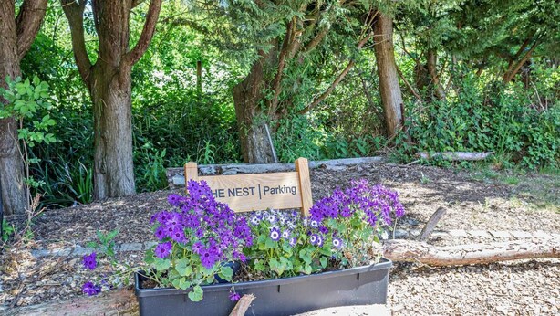 Un panneau en bois portant l’inscription « The Nest | Parking », entouré de fleurs violettes et de verdure.