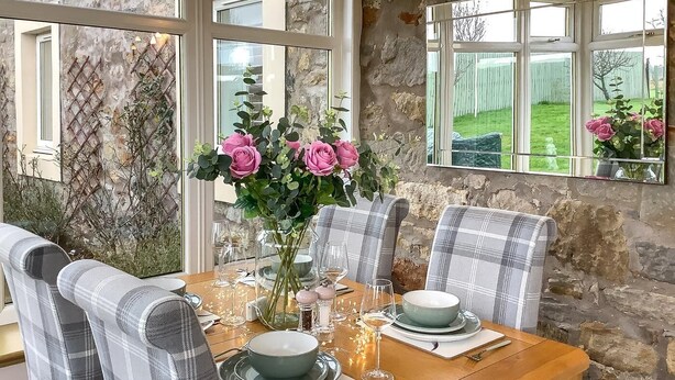 Une table à manger, entourée de chaises, agrémentée d’un vase de fleurs, et offrant une vue sur un jardin grâce à de grandes fenêtres.
