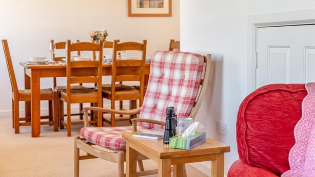 A dining area with wooden chairs and a table, a red armchair, and a small table with a magazine and a thermos. - 3 Bed in Polruan (oc-g30535) (Fowey)