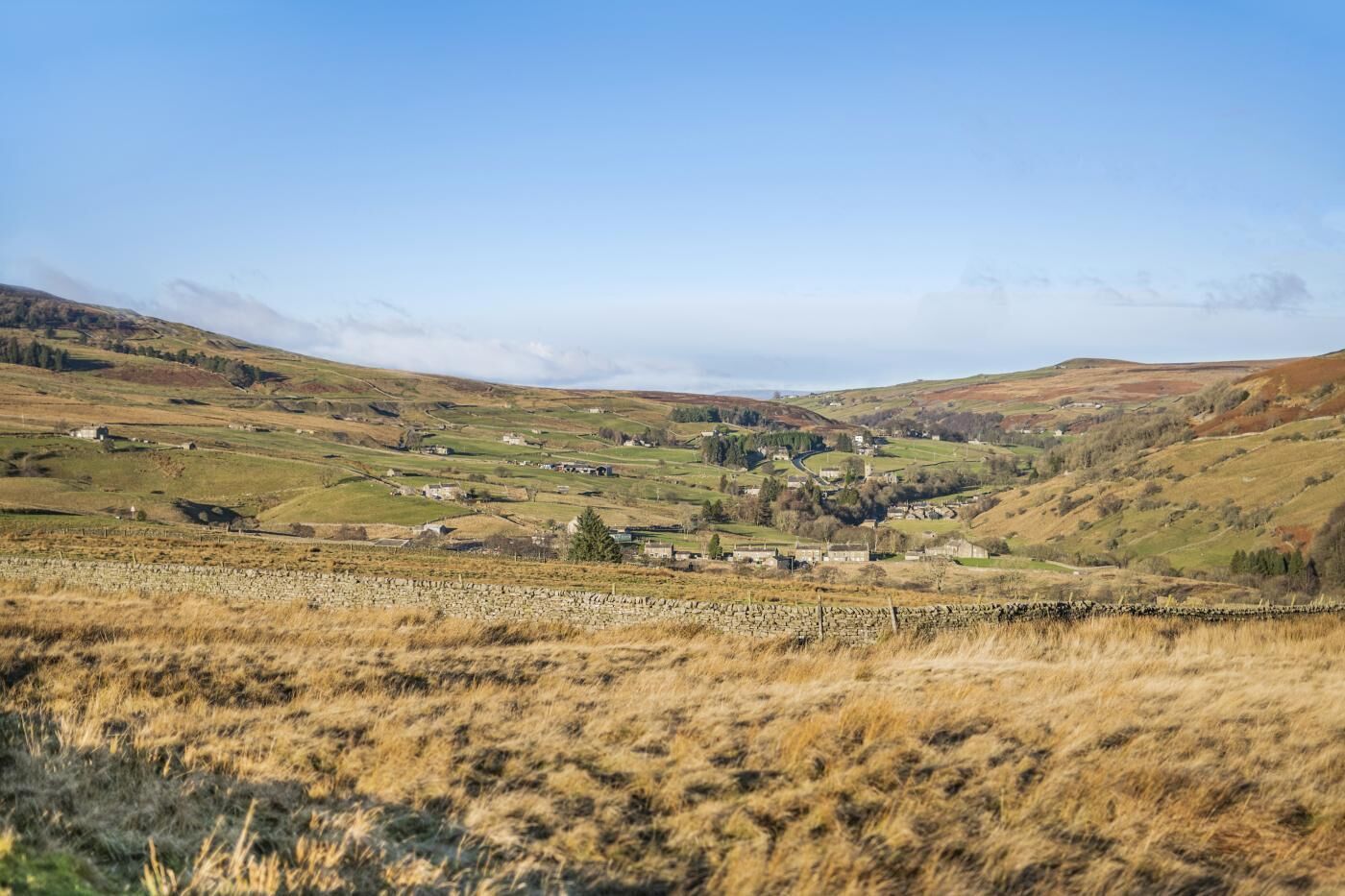 A rural landscape with rolling hills, a stone wall in the foreground, and a small village with buildings and a church in the distance.