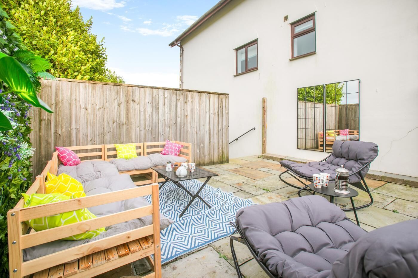 A patio area with a wooden bench, a cushioned chair, and a small table with cups and a teapot.