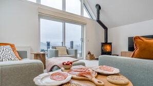 Ein modernes Wohnzimmer mit großem Fenster, einer Couch, einem Tisch mit Speisen und einem Kaminofen.