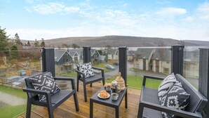 Ein Balkon mit drei schwarzen Stühlen, jeder mit einem Kissen in geometrischem Muster, einem kleinen Tisch mit einem Croissant und einer Tasse, sowie einer Laterne. Der Balkon blickt auf eine Wohngegend mit Häusern und einer Hügelkette in der Ferne.