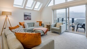 Ein geräumiges Wohnzimmer mit einer großen Couch, Sesseln und einem Couchtisch. Mehrere Fenster sorgen für natürliches Licht und bieten einen Ausblick ins Freie.