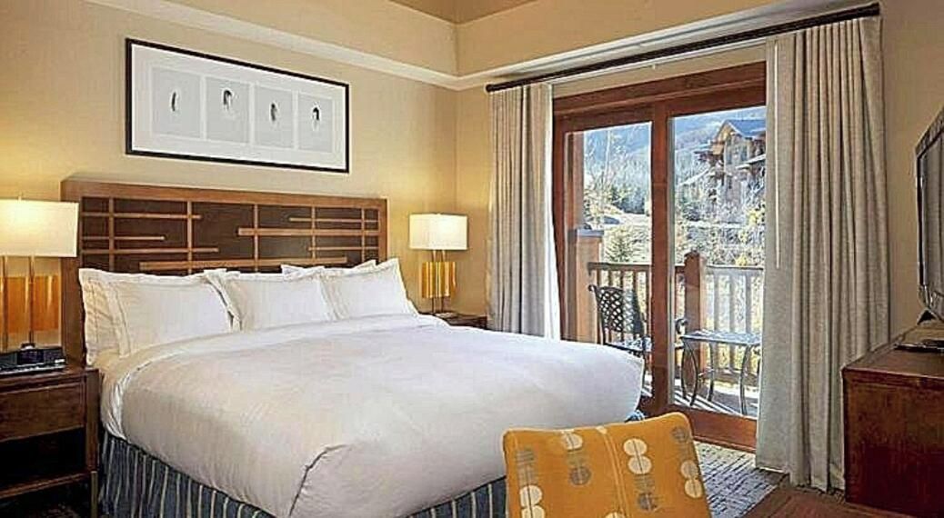 A hotel room with a large bed, two bedside lamps, a wooden headboard, a framed picture above the bed, and a balcony with curtains.