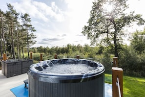 A hot tub surrounded by a deck, with a pool and trees in the background.