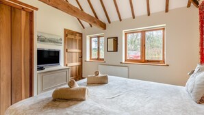 Ein Schlafzimmer mit einem großen Bett, Holzbalken, einem Fernseher und Fenstern mit Blick auf Grünfläche.