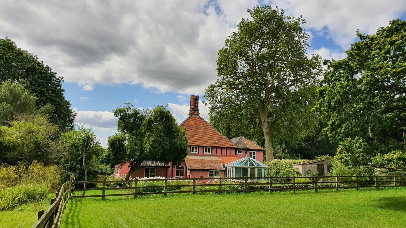 Une maison rouge avec un toit vert, entourée d’une végétation luxuriante et d’une clôture en bois.
