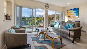 A spacious living room with a large sliding glass door leading to a balcony, a sofa set, a coffee table, and a clock on the wall.
