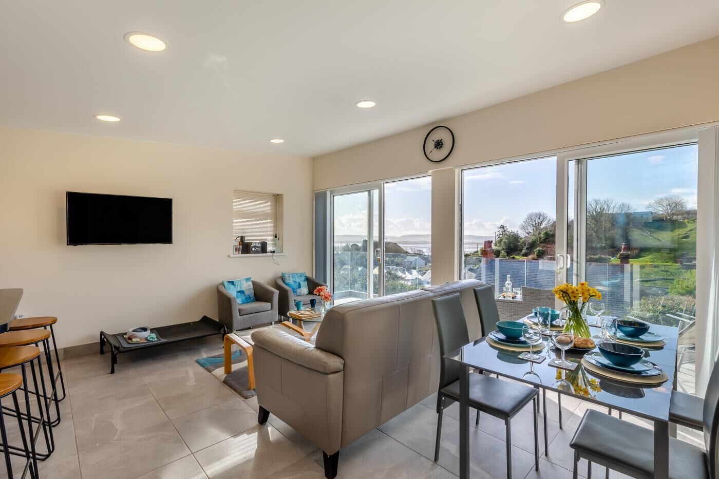 A modern living room with a dining area, large windows, a flat-screen TV, and a view of the city.