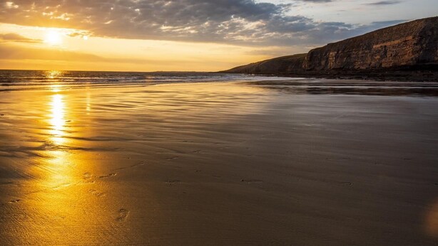 Une plage au coucher du soleil, avec des vagues calmes et une falaise rocheuse.