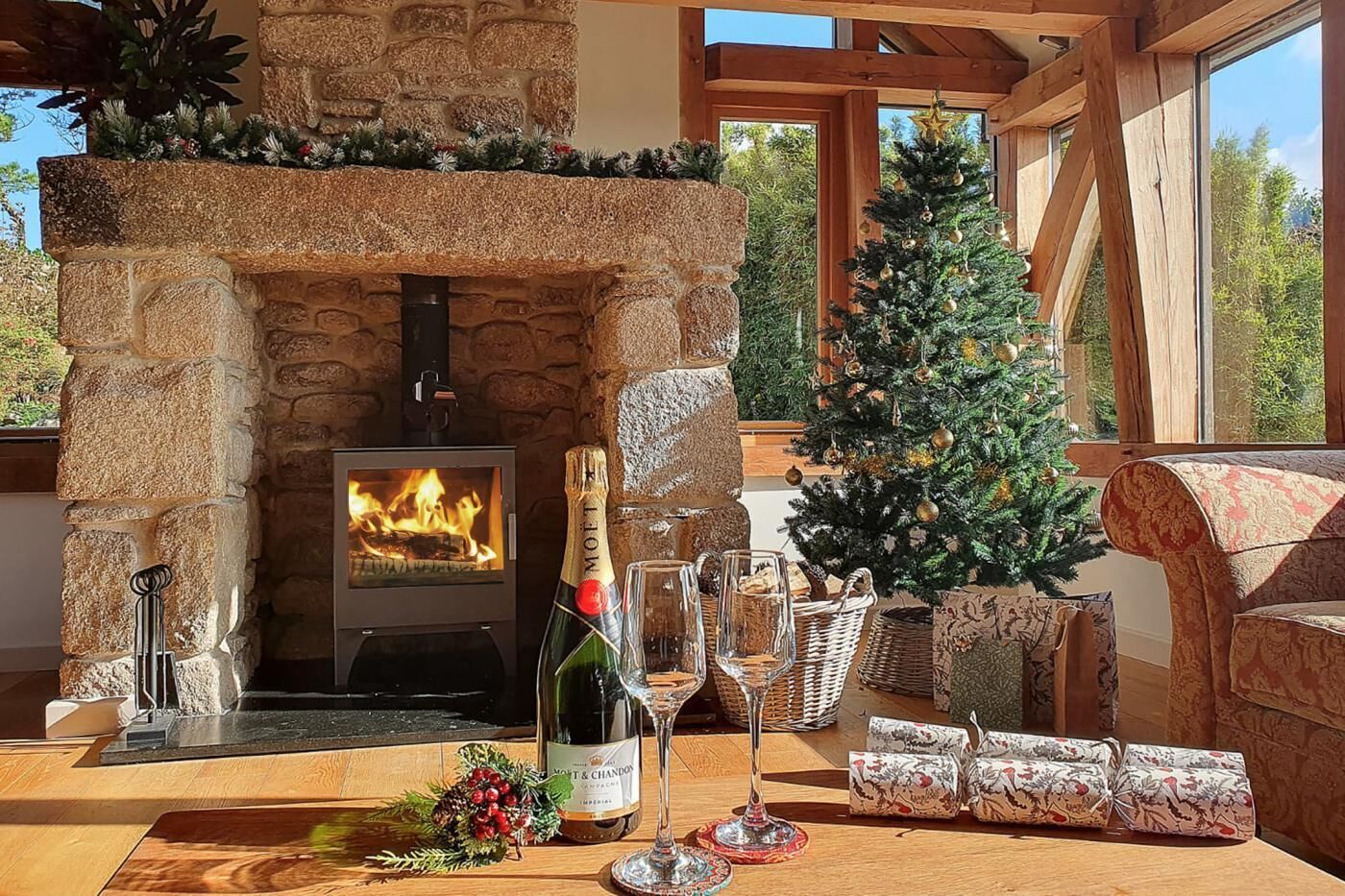 A cozy fireplace with a stone mantle, a bottle of champagne, and a Christmas tree in a well-lit room with wooden beams and large windows.