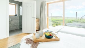Une chambre avec un grand lit, une table de chevet en bois sur laquelle se trouvent une assiette de fruits et des tasses à café, une salle de bain avec une douche en verre, et une vue sur la verdure à l’extérieur.