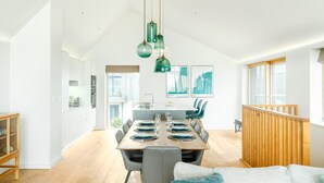 Un vaste espace de salle à manger avec une longue table dressée pour quatre personnes, une cuisine en arrière-plan et une vue sur l’extérieur.