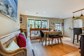 A cozy dining area with a wooden table, chairs, and a bench. There is a window with green shutters and a view of trees outside. - 2 Bed in St. Mawgan (oc-l30651) (Saint Mawgan)