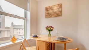 Un coin repas avec une table ronde en bois, deux chaises, un vase de fleurs et une fenêtre donnant sur des immeubles.