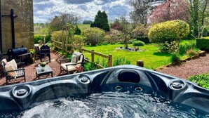 A hot tub in a garden with a wooden deck, chairs, and a barbecue. - 5 Bed in Biddlestone (oc-b30105) (Biddlestone)