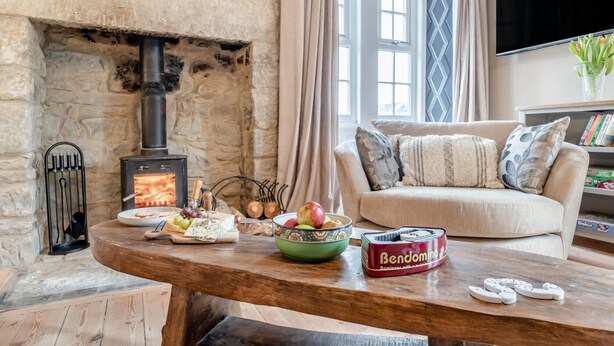 A cozy living room with a stone fireplace, a comfortable sofa, a wooden coffee table with snacks, and a television. - 5 Bed in Biddlestone (oc-b30105) (Biddlestone)