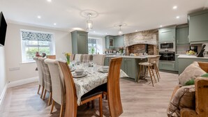 A modern kitchen with a central island, built-in appliances, and a dining area with wooden chairs and a table set for six. - 5 Bed in Biddlestone (oc-b30105) (Biddlestone)