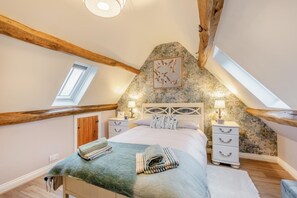 Ein Schlafzimmer mit schrägen Wänden, Holzbalken, einem Bett mit Kopfteil, Nachttischen, einem Wandbild und einem Dachfenster.