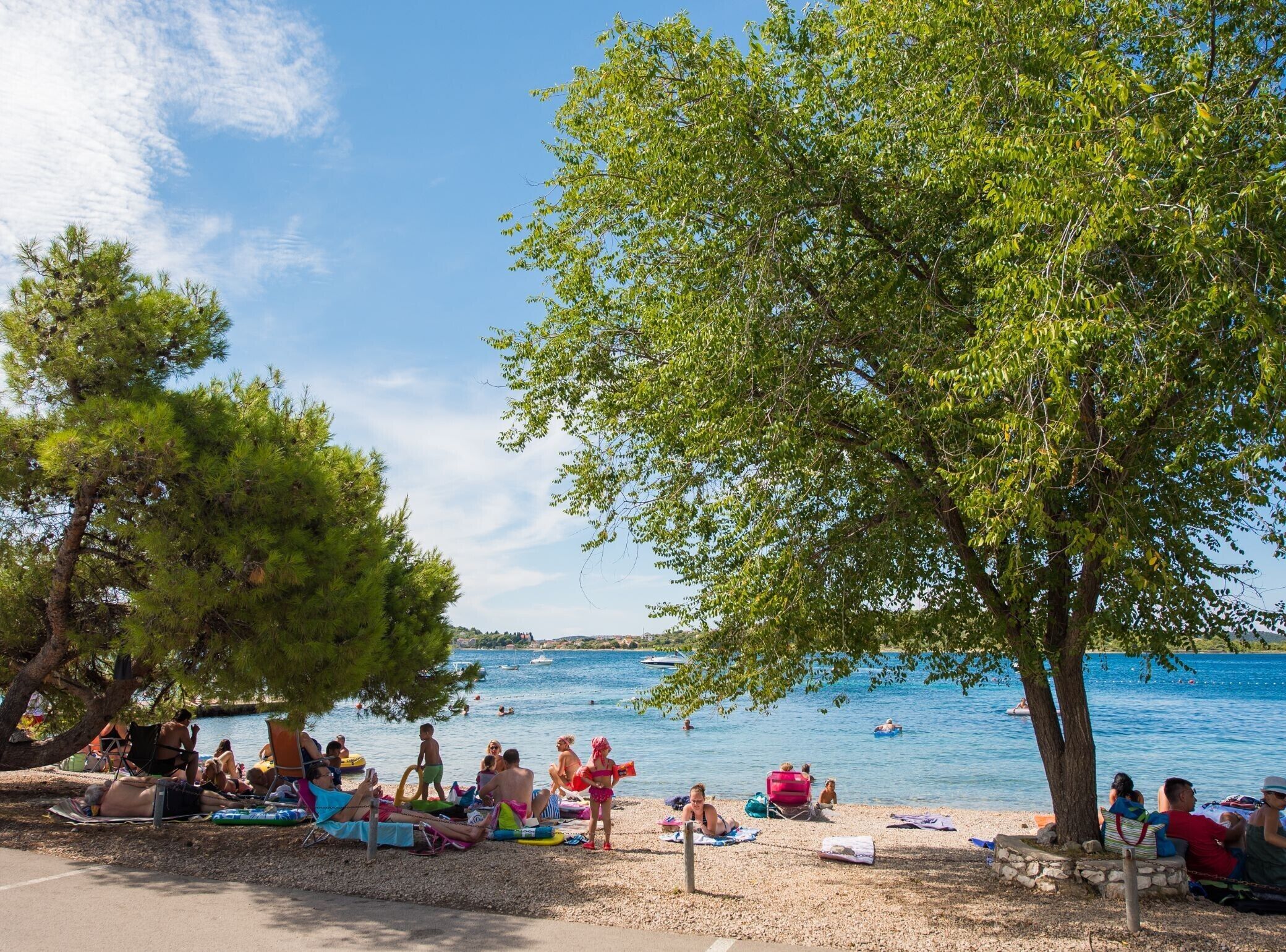 Ein Strand mit badenden und sonnenden Menschen, umgeben von Bäumen und blauem Himmel.