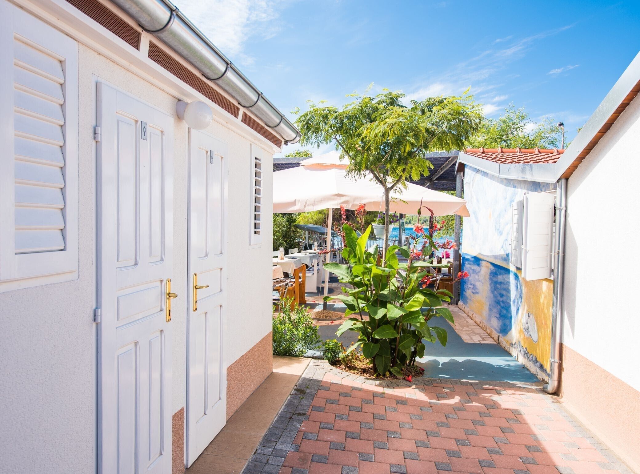 A narrow alleyway with a white door on the left, a small garden with a tree and plants, and a mural on the right wall.