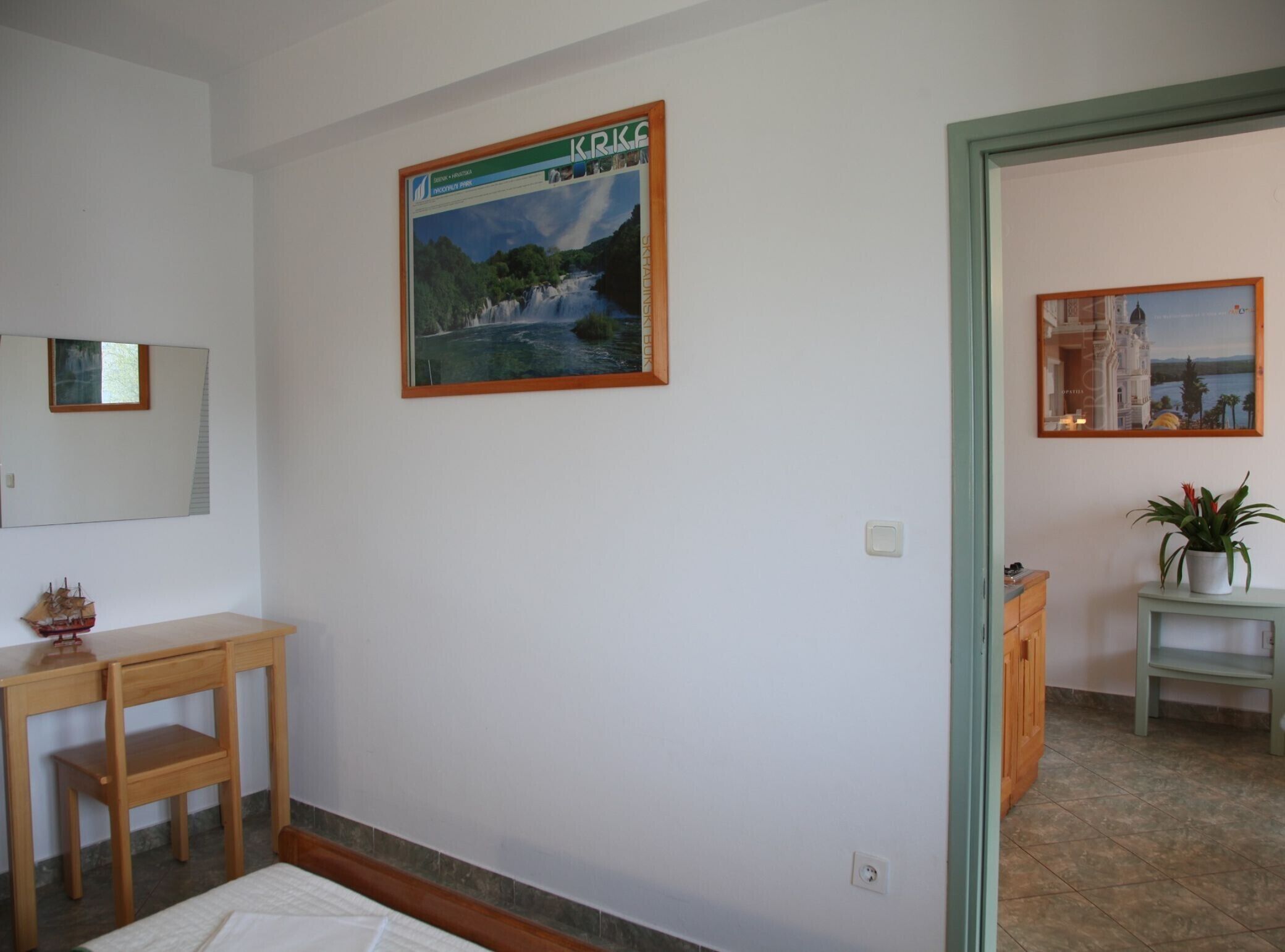 Une chambre à coucher comprenant un lit, un bureau en bois, une petite table, un miroir et un tableau encadré représentant une cascade.