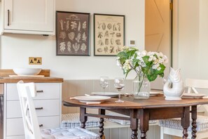 A dining area with a wooden table set for four, white chairs, a vase with flowers, and framed botanical prints on the wall. - 2 Bed in Llangedwyn (oc-l30530) (Llangedwyn)