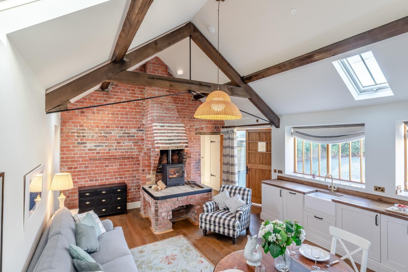 A loft-style living space with exposed wooden beams, a brick fireplace, a dining area, and a kitchen with a large window.