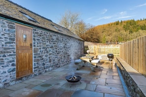 A stone building with a wooden door, a fire pit, outdoor seating, and a view of a forested hillside. - 2 Bed in Llangedwyn (oc-l30530) (Llangedwyn)