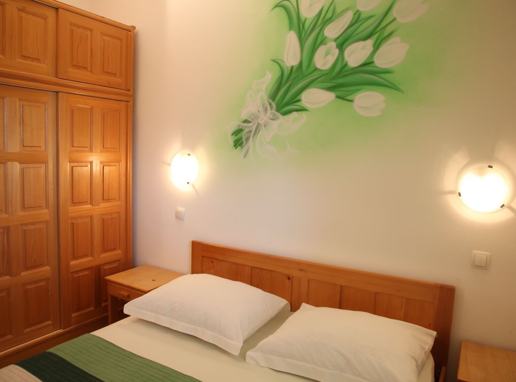 A bedroom with a wooden headboard, two bedside tables, a bed with white bedding, and a wall with a floral mural.