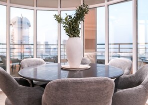 A round table with a white vase and a plant in the center, surrounded by grey upholstered chairs, in a room with large windows offering a view of the city. - Stayshort - 2 Bedroom in Dubai Marina With Huge Balcony (Dubai)