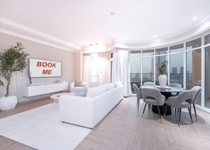 A modern living room with a white sofa, a dining table with chairs, a large window with curtains, and a 'BOOK ME' sign on the wall. - Stayshort - 2 Bedroom in Dubai Marina With Huge Balcony (Dubai)