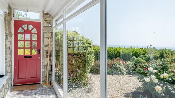 A red door with a glass panel, a stone wall, and a view of a garden with various plants and flowers. - 2 Bed in Ventnor (oc-w30525) (Ventnor)