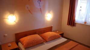 Een slaapkamer met een houten hoofdbord, twee oranje kussens, een nachtkastje met een lamp en een raam met gordijnen.